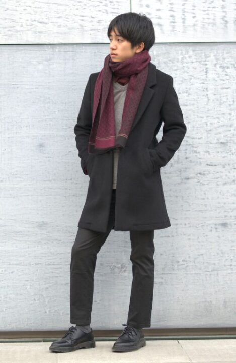 グレーのニットと黒のチェスターコートを着用し、首に赤いストールを巻いた男性