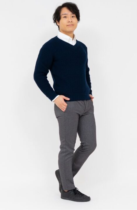 白いシャツの上にVネックのネイビーのニットを着用し、グレーのパンツを履いた男性